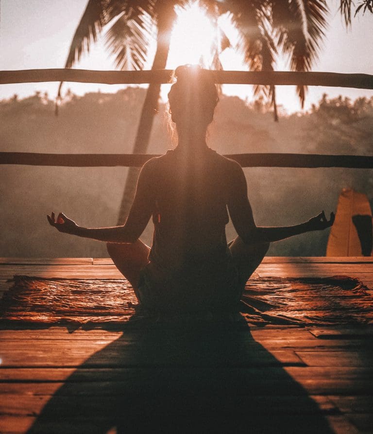 Femme de dos assise en position du lotus face au soleil couchant, sur une terrasse en bois face à un palmier.