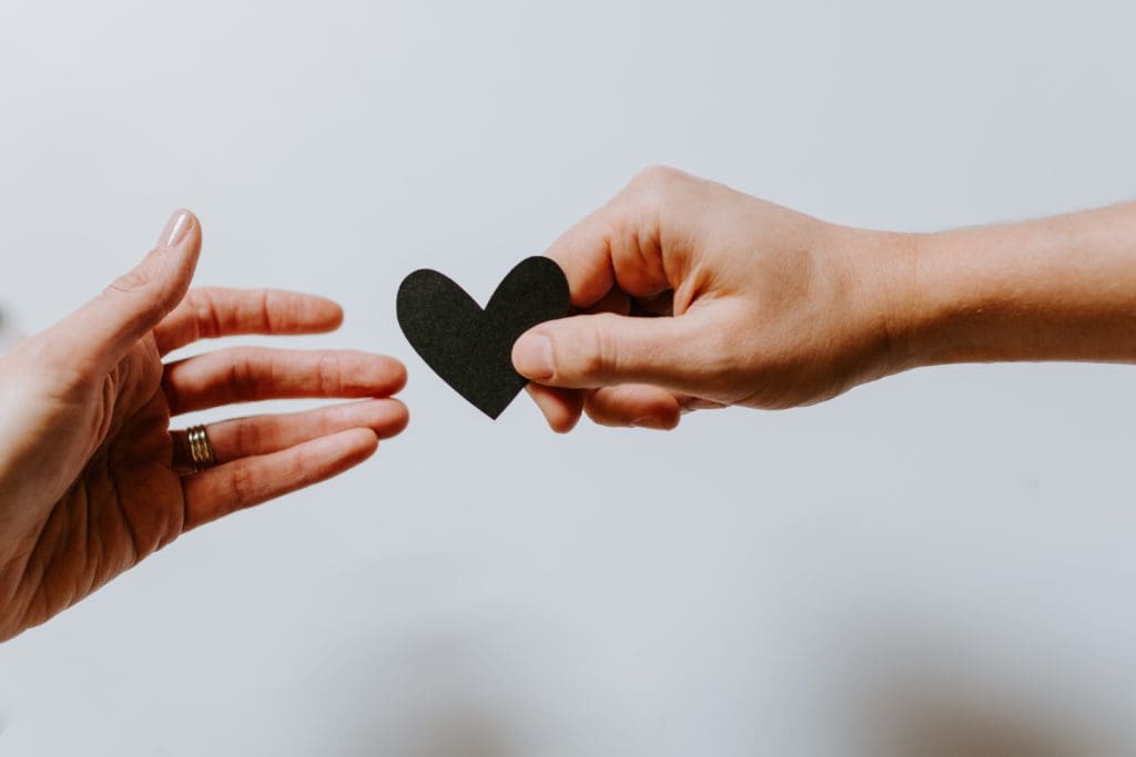 Une main tend un coeur en papier noir à une autre main sur la gauche.