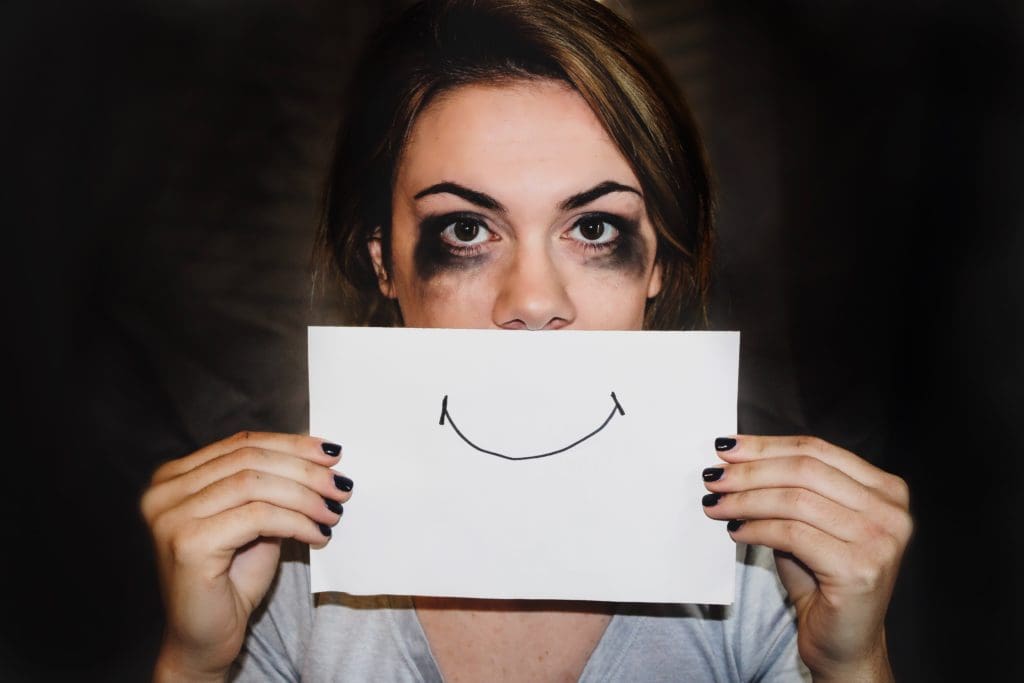 Une femme au visage maculé de maquillage qui a coulé (yeux charbonneux et joues noircies) couvre sa bouche avec une feuille sur laquelle est dessiné un sourire très simplifié au stylo.