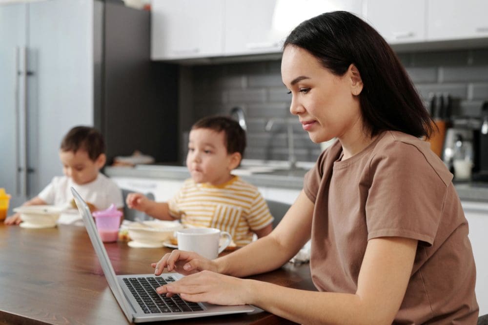a woman in brown shirt typing on her laptop while sitting beside her kids